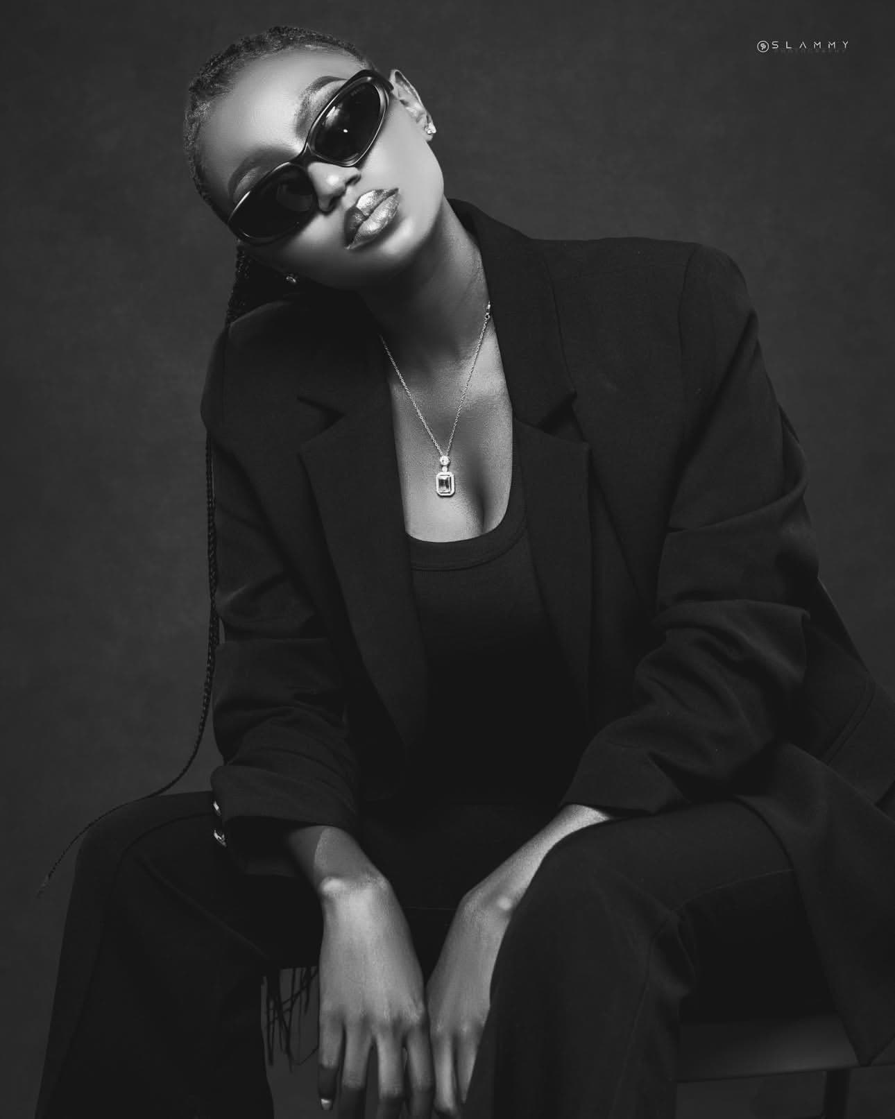 Black and white studio portrait of Abiola Ayeni seated in a black blazer, sunglasses and a diamond pendant necklace