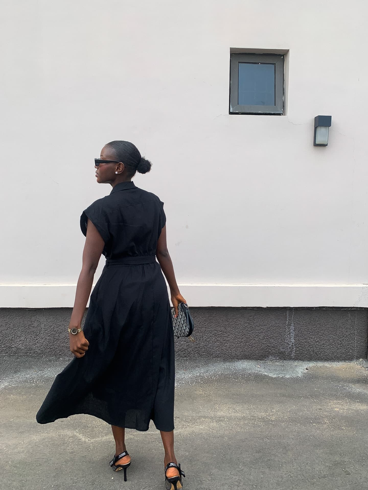 Abiola Ayeni walking away from camera in a black midi shirt dress and sunglasses, against a plain white wall