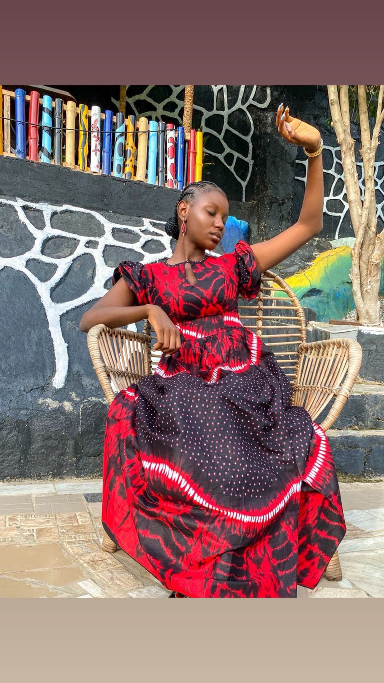Abiola Ayeni seated in a red and black tie-dye maxi dress on a woven rattan chair, against a hand-painted mural wall